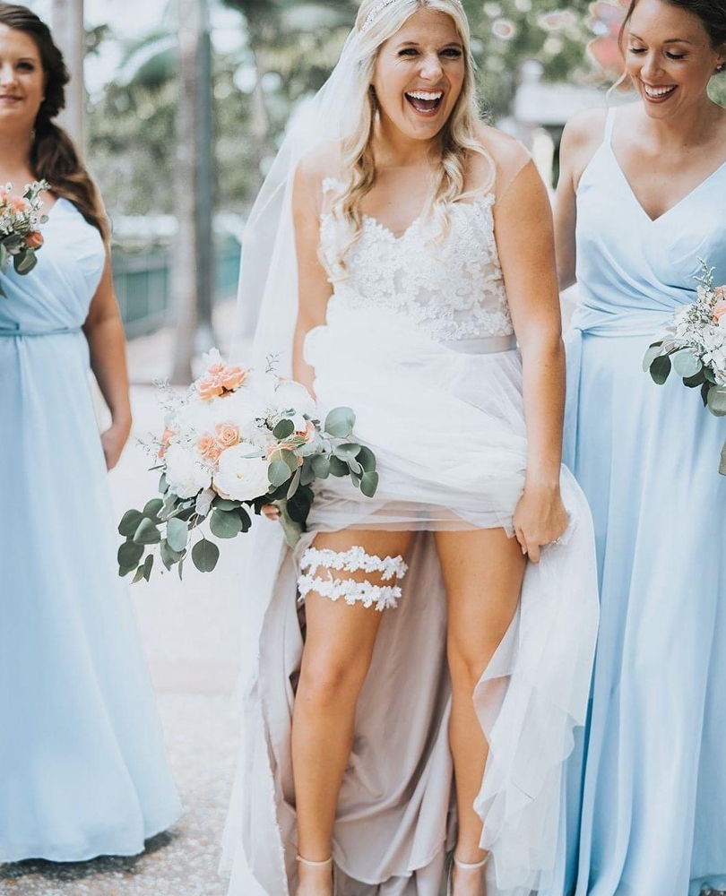 Bride lifts her lace dress to reveal a floral garter while smiling, flanked by bridesmaids in pale blue gowns holding bouquets outdoors.