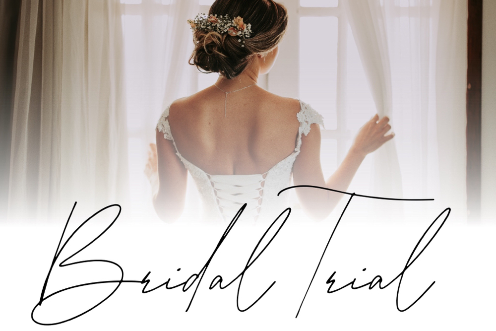 Bride in an open-back lace wedding gown seen from behind, standing by a sunlit window with 'Bridal Trial' cursive text across the bottom.